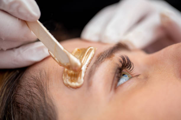 Beautician Applying Wax with Wooden Spatula Above Woman's Eyebrow